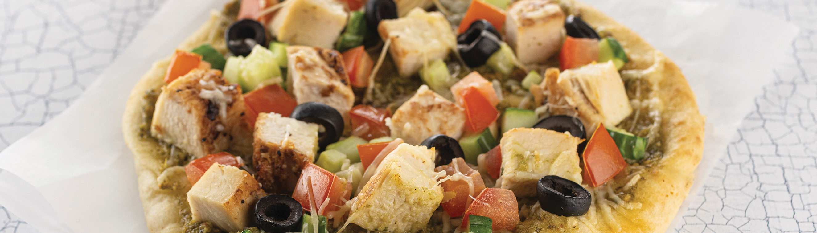 A Crispy pita topped with pesto, roasted chicken, shredded Parmesan, tomatoes, olives and English cucumbers with Italian cucumber salad.