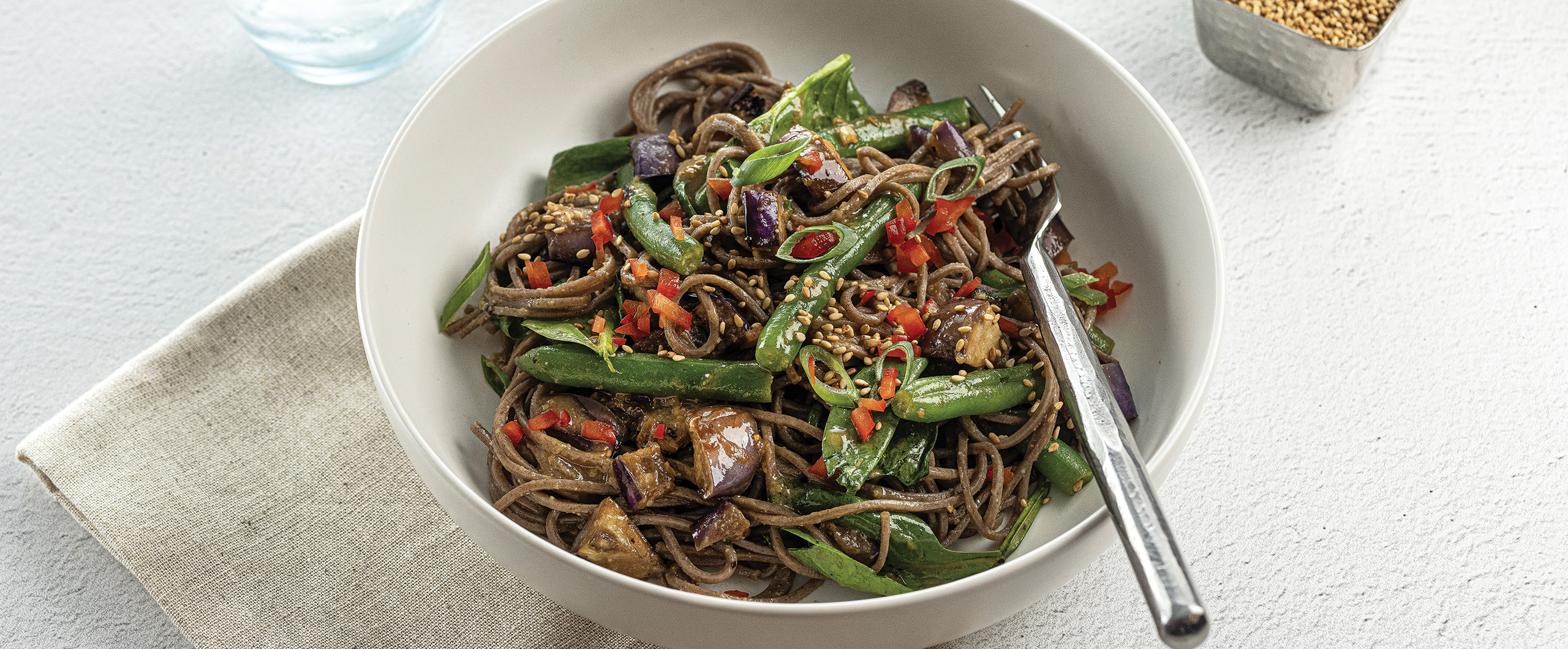 A large white bowl filled with Miso-glazed eggplant and green beans with soba noodles, fresh baby spinach, and sesame seeds garnished with green onions and red peppers.