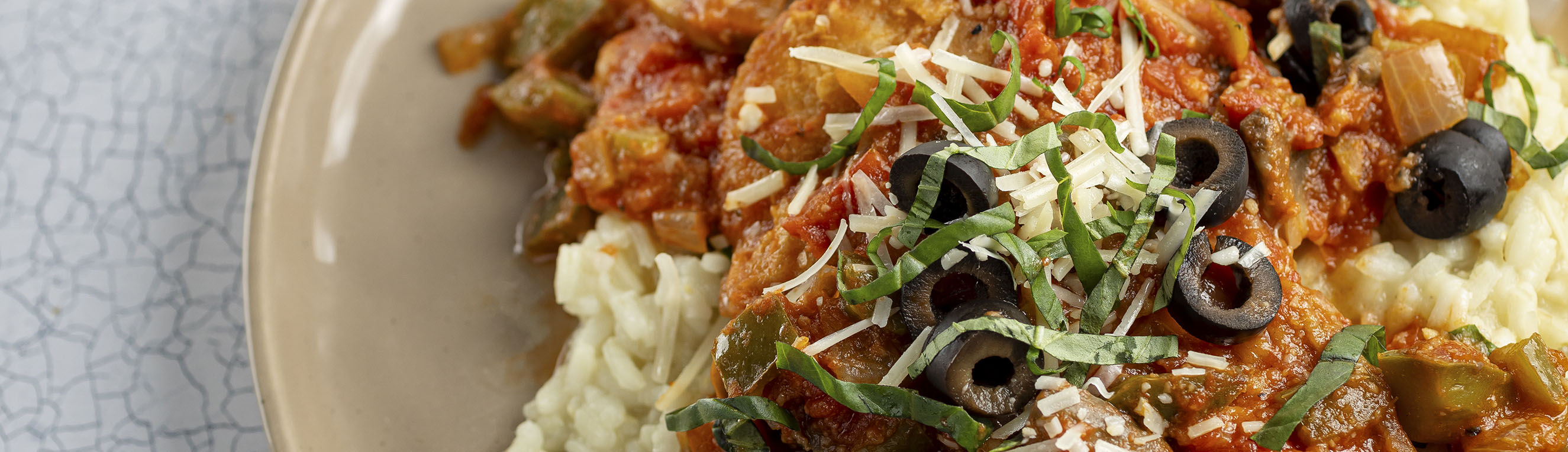 An entree plate featuring chicken thighs in tomato sauce over risotto, topped with black olives, Parmesan cheese and fresh basil